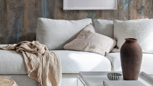 Eine graue Couch mit einer Decke in beige, einem Kissen in rose und einem Kissen in Strick-Optik. Im Vordergrund ist eine betagte Vase zu sehen