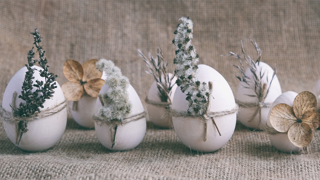 Sieben weiße Ostereier in einer Reihe aufgestellt, verziert mit unterschiedlichen und meist getrockneten Blüten oder kleinen Ästen. 