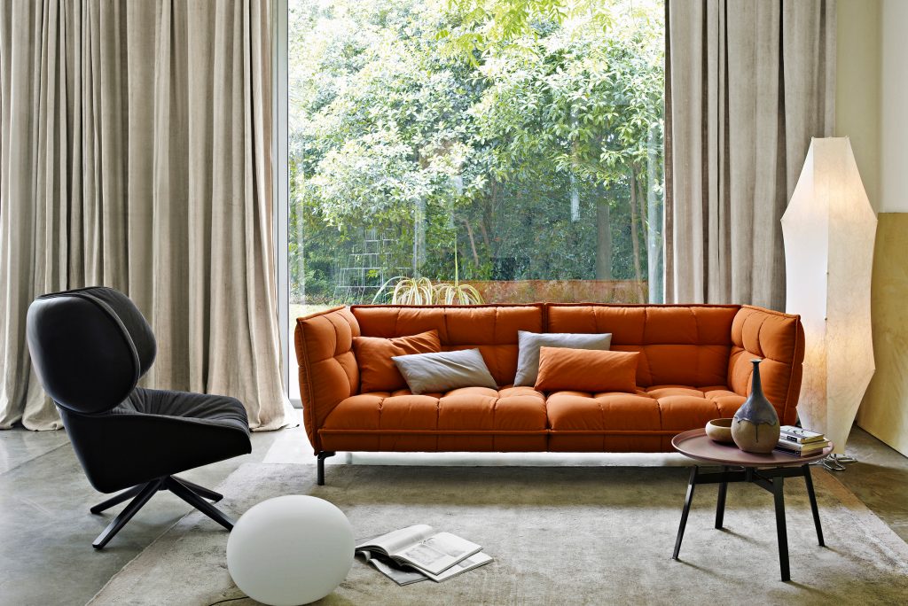 Eine orange Couch mit bunten Kissen steht vor einem großen Fenster. Blick in den Garten. 