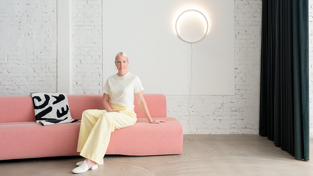 Designerin Sabine Marcelis sitzt auf einer pinken Couch vor einer weißen Wand. An dieser Wand befindet sich eine der neuen Leuchten